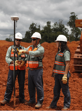 Photo of two men and a woman talking and observing equipment that resembles an iron bar in a operation area. They are wearing green button-down Vale shirt, orange vests with fluorescent stripes, jeans, gloves, boots, goggles and helmets.