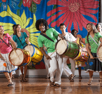 Jovens tocam tambores e sorriem, como se estivessem cantando. Atrás há um tecido estampado com as cores azul, vermelho, amarelo e verde.