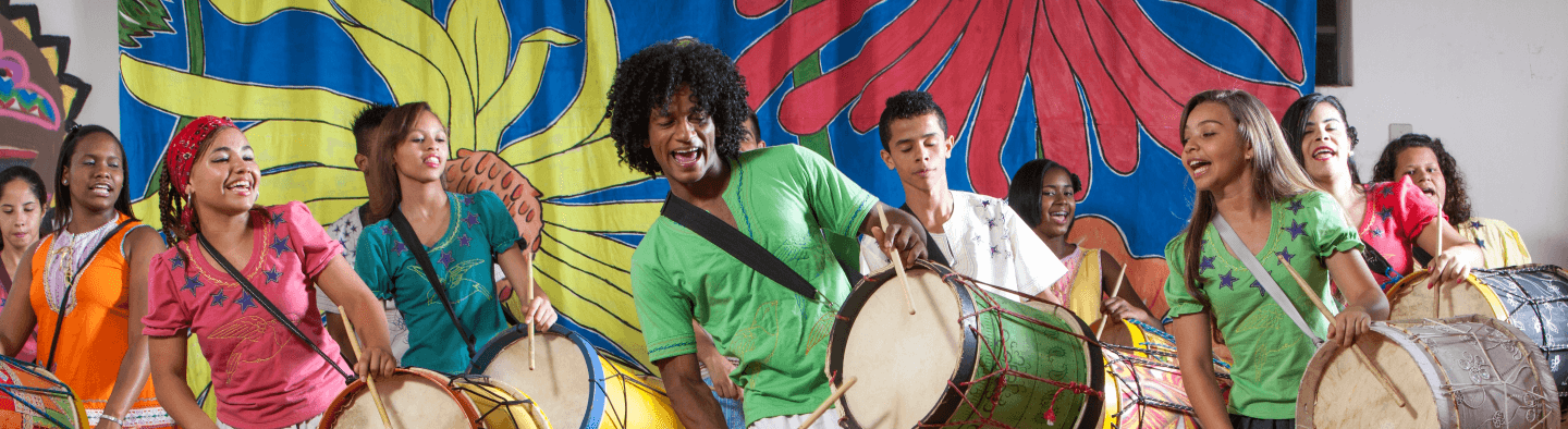 Jovens tocam tambores e sorriem, como se estivessem cantando. Atrás há um tecido estampado com as cores azul, vermelho, amarelo e verde.