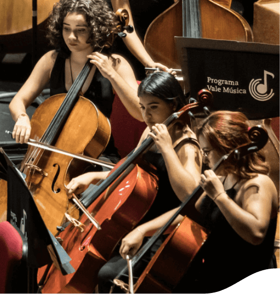 Orquestra vestida de preto. É possível ver três mulheres, todas tocando violoncelo, e um cartaz escrito “Programa Vale Música”