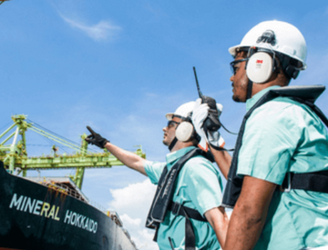 Two Vale employees facing a ship in an operational area. The two are wearing green shirts, protection equipment fixed to the body, goggles, ear muffs, and white helmets.