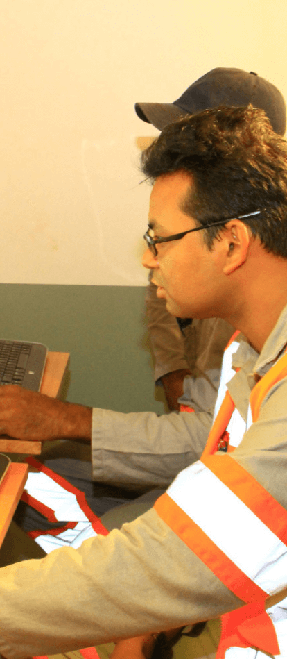 Foto de um homem mexendo no computador e duas pessoas ao lado dele. Ele está usando camisa cinza de botões, colete laranja com listras fluorescentes e óculos de grau.