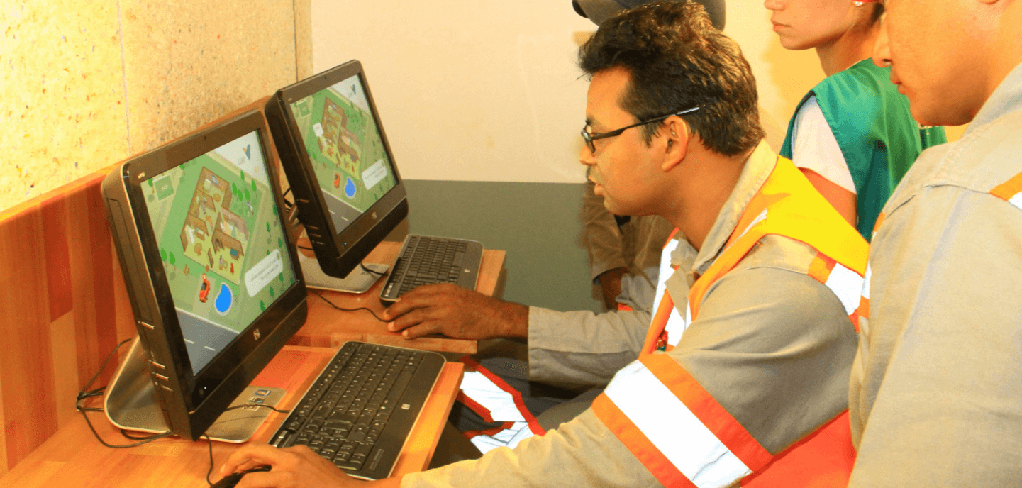 Foto de um homem mexendo no computador e duas pessoas ao lado dele. Ele está usando camisa cinza de botões, colete laranja com listras fluorescentes e óculos de grau.