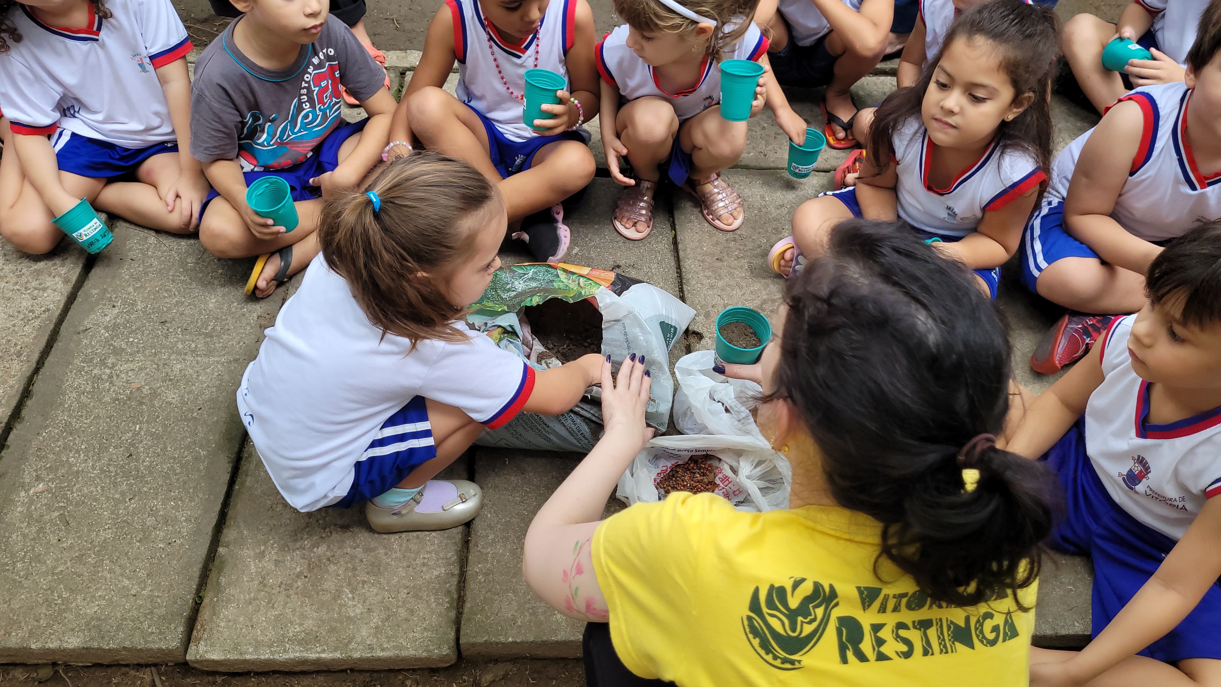 Uma mulher está reunida com diversas crianças, que vestem uniformes escolares. Elas estão mexendo em um saco com terra.