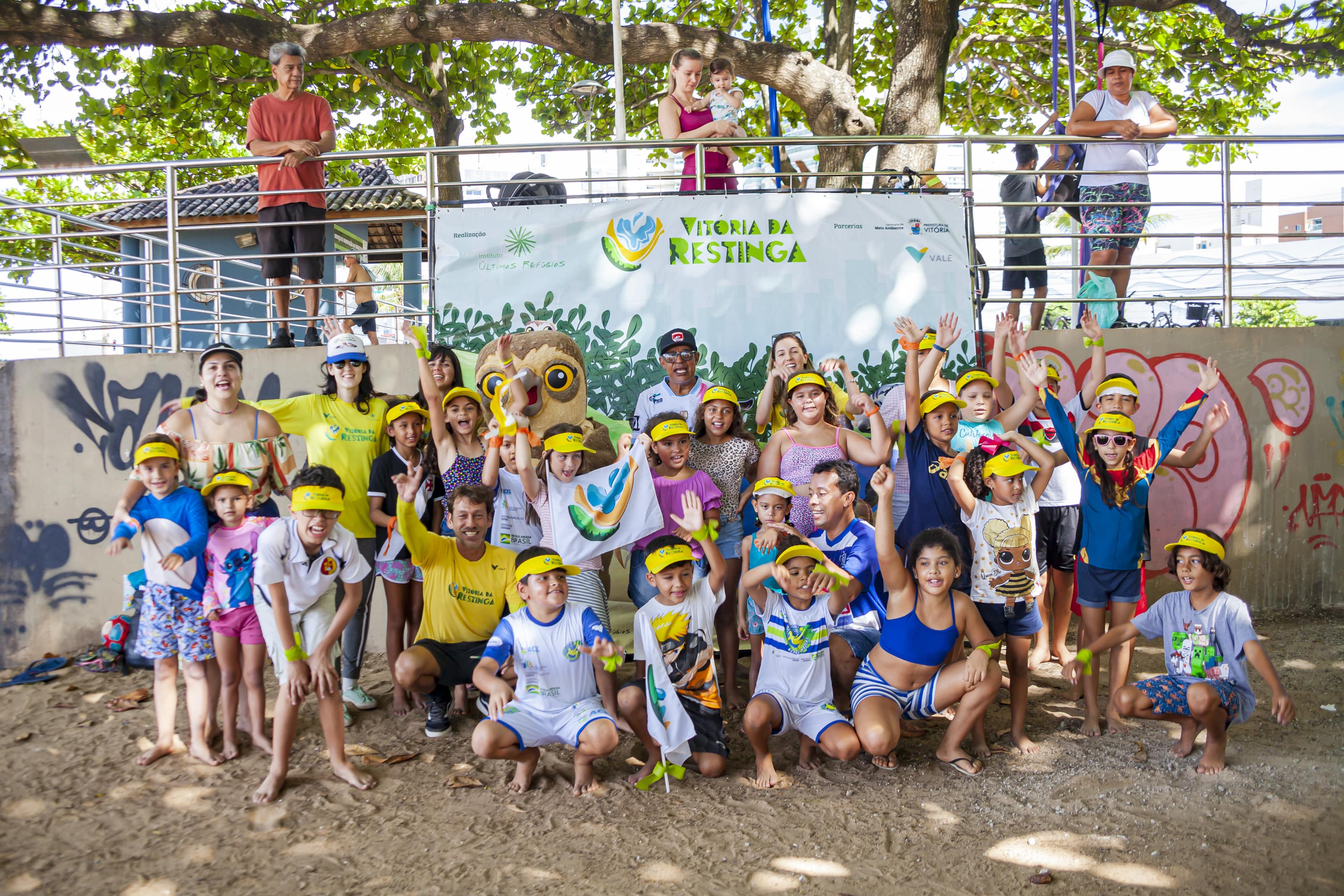 Diversas crianças e alguns adultos estão reunidas, junto de um mascote vestido de coruja. Elas estão com bonés amarelos e ao fundo há uma placa “Vitória da Restinga”