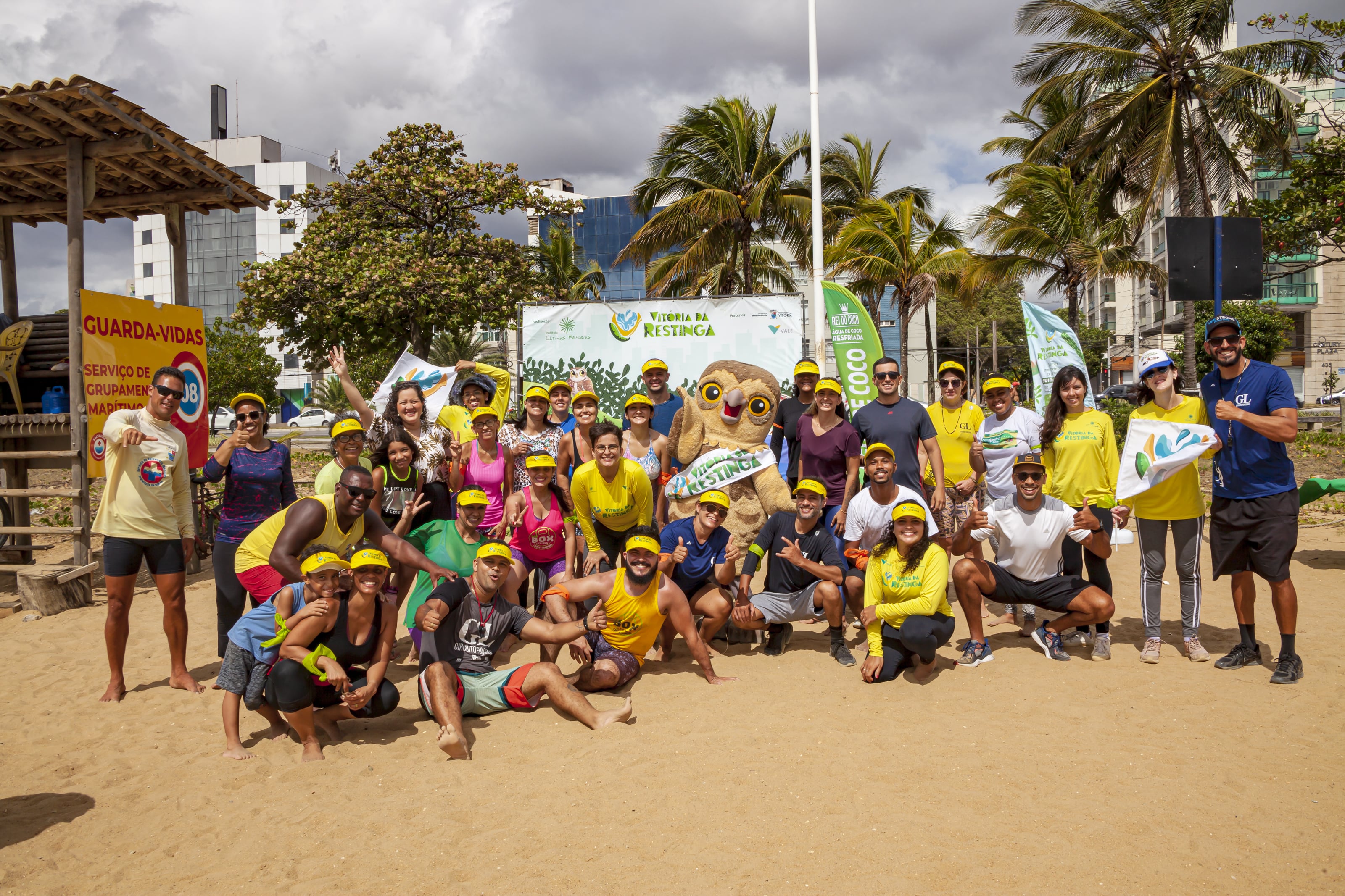 Diversas pessoas estão reunidas na areia da praia, junto de um mascote vestido de coruja. Elas estão com bonés amarelos e ao fundo há uma placa “Vitória da Restinga”