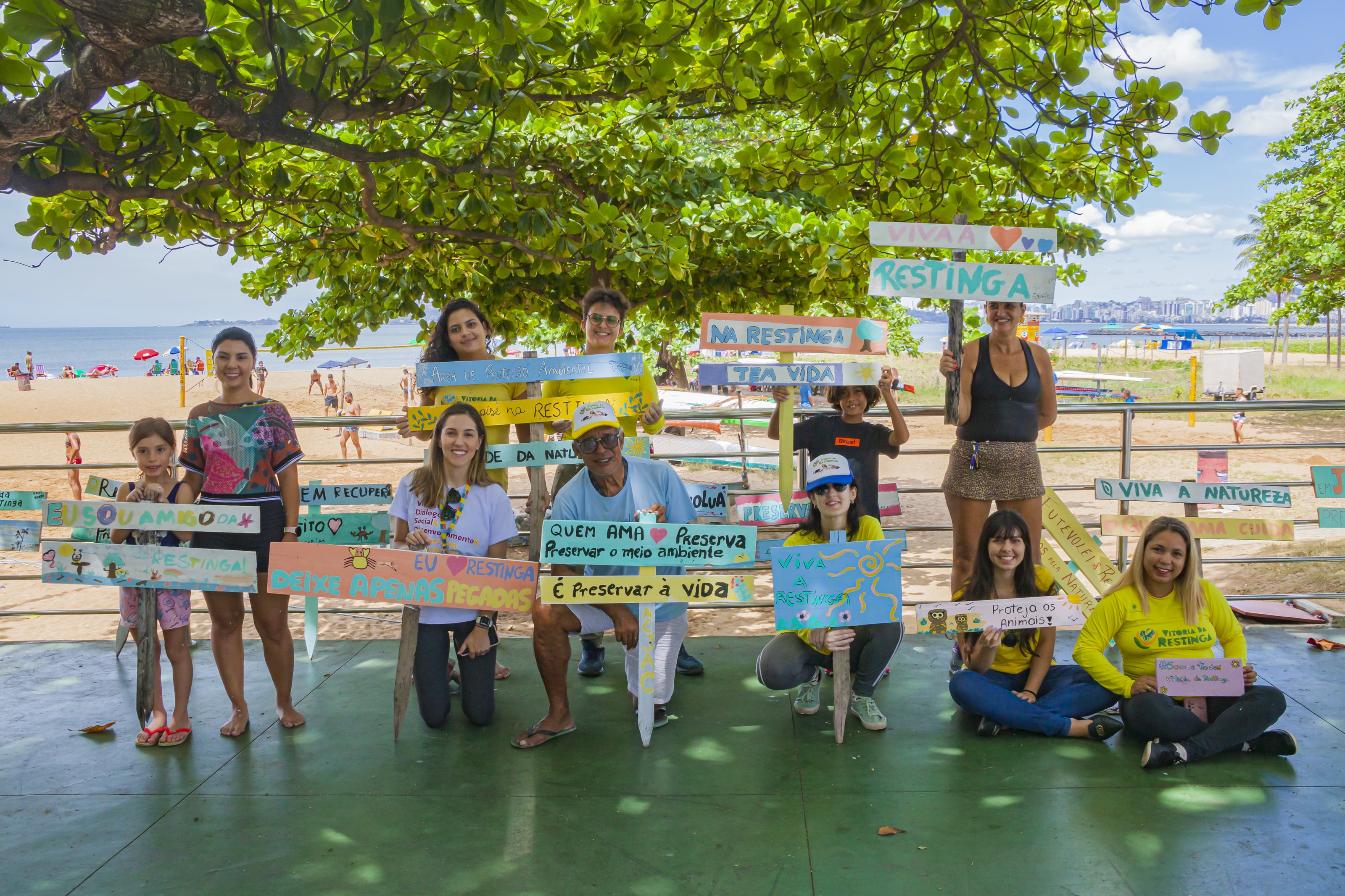 Crianças e adultos posam para foto segurando placas sobre a preservação da restinga. Ao fundo é possível ver a praia