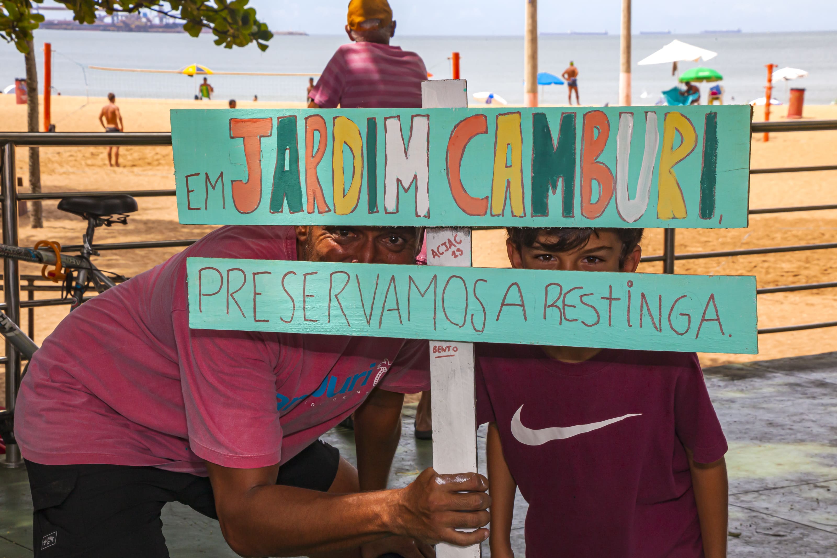Um homem e um menino posam para foto atrás de uma placa em que está escrito “Em jardim Camburi preservamos a restinga”