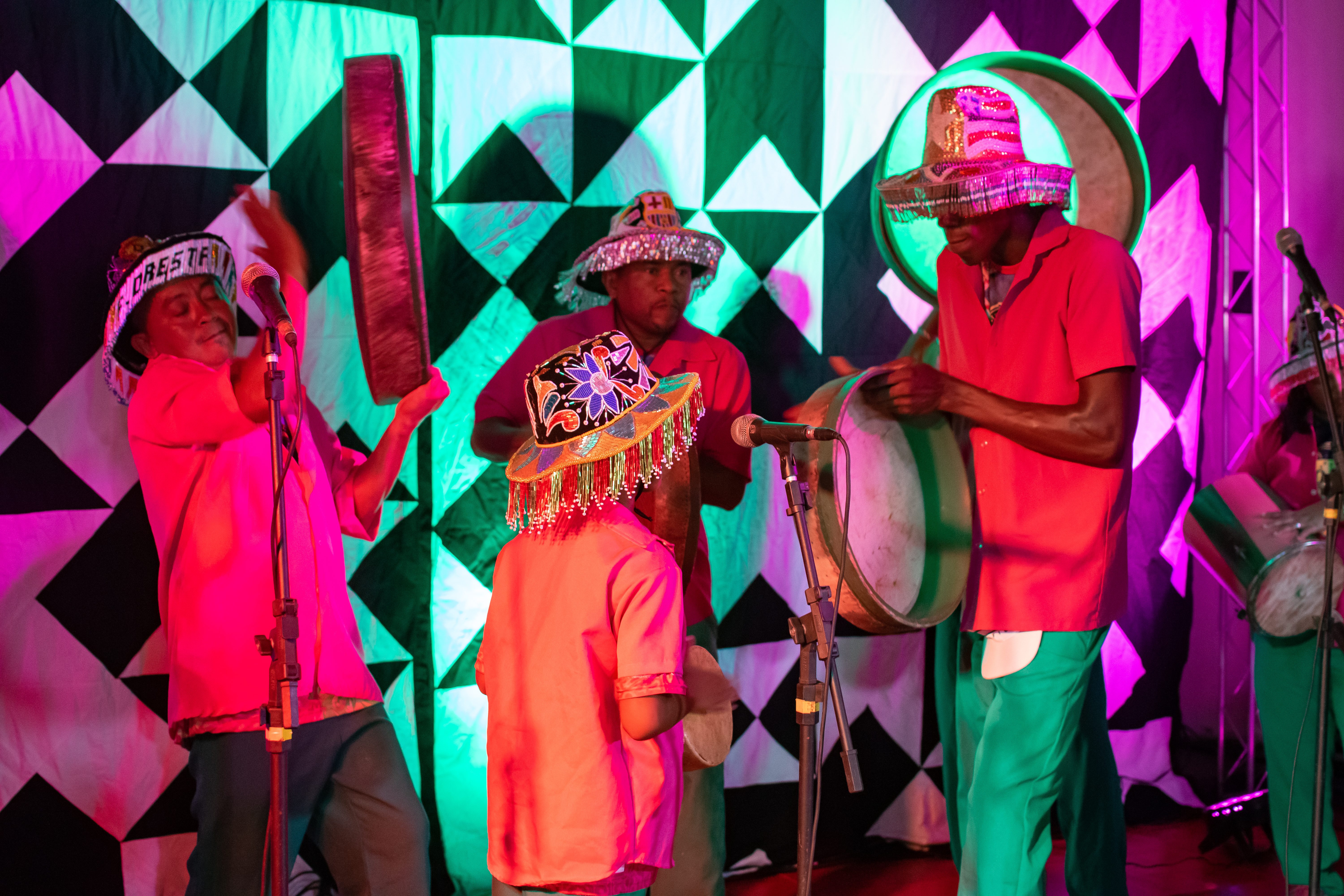 Foto de três homens e uma criança em cima de um palco com instrumentos musicais e microfone. Todos estão usando chapéu colorido