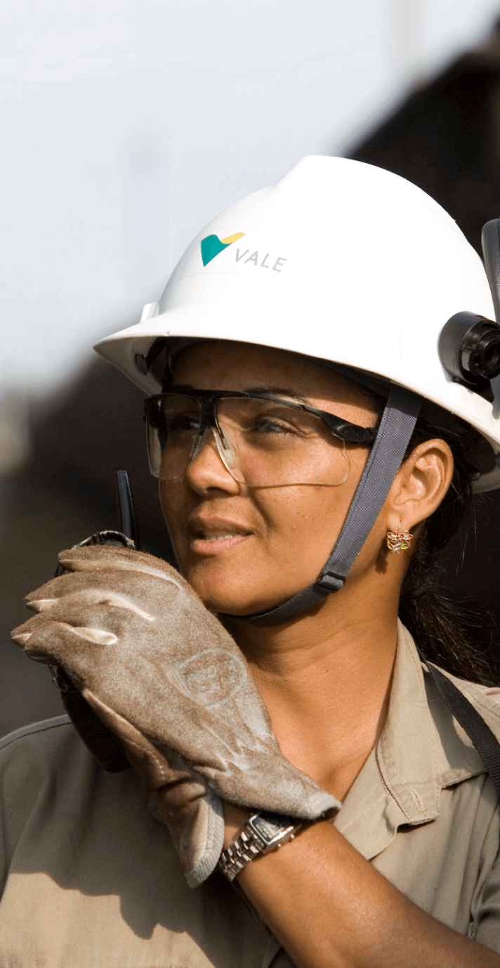 Vale employee in an outdoor area. She looks to the horizon while holding a radio transmitter close to her mouth. She is wearing a helmet, gloves and goggles.