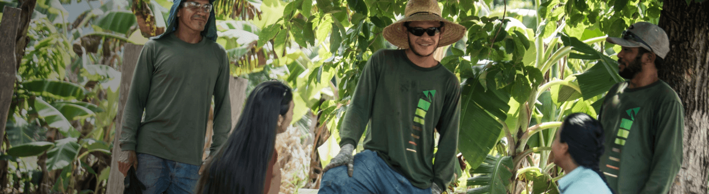 Em uma área de vegetação, três homens e duas mulheres conversam. Os homens estão de camiseta verde escura de manga longa, proteção contra o sol na cabeça - como chapéu e boné - e usam óculos e luva.