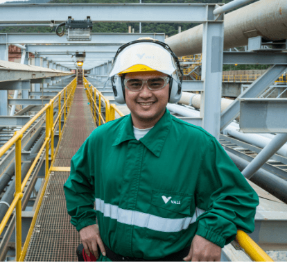 A man is smiling on a footbridge in an operation area. He is wearing a dark green shirt, white helmet, goggles, and ear protection.