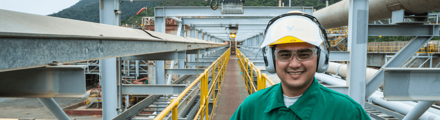 Um homem sorrindo em uma passarela de uma área de operação. Ele usa camisa verde escura, capacete branco, óculos de proteção e proteção nos ouvidos.