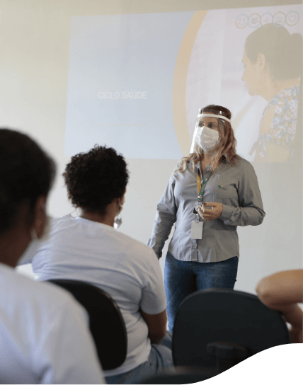 Mulher com camisa cinza, calça jeans, crachá e duas máscaras faciais fazendo uma apresentação com o apoio de um telão, que está atrás dela. Na frente dela, há algumas pessoas sentadas assistindo o que está sendo mostrado no telão.