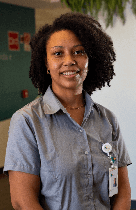 Mulher negra sorrindo em um escritório. Ela está com os cabelos soltos, usa uma camisa cinza e crachá.