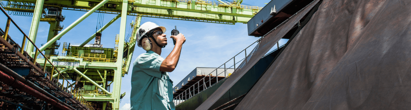 Empregado da Vale em uma área operacional. Ele está usando camisa verde clara, óculos de proteção, protetores de ouvido e capacete branco, além de estar segurando um rádio comunicador em uma das mãos.