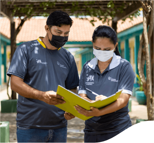 A man and a woman standing side by side writing in a yellow notebook they both hold. The two are wearing jeans, gray shirts and face masks.