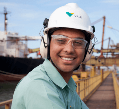 Homem sorrindo em um espaço de operações com uma estrutura de metal e um navio ao fundo. Ele usa camisa verde clara, óculos de proteção, protetores de ouvido e capacete branco.