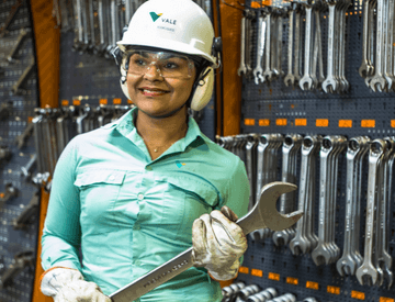 A woman standing, holding a steel tool in a space full of other tools. She is wearing light green uniform, gloves, goggles, ear muffs and a white helmet with Vale logo.