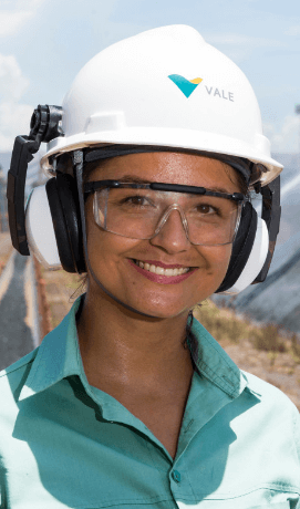 Foto tirada de rosto de uma empregada da Vale sorrindo em uma área operacional. Ela usa camisa verde clara, óculos de proteção, protetores de ouvido e capacete branco com logotipo da Vale.