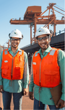 Two men smiling in an operational space. The two wear green uniform shirts, orange vests, goggles and white helmets with Vale logo. One of them holds a clipboard in his hands.