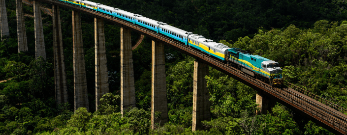 Trem Vale seguindo por uma ferrovia elevada. Em volta é possível ver uma paisagem montanhosa e repleta de vegetação.