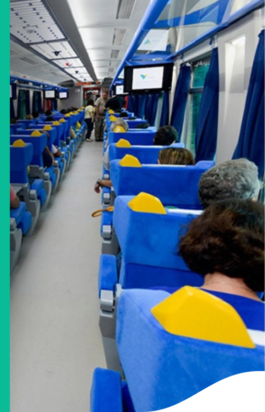 Image of train seats with passengers seated. There are two people standing, circulating in the corridor. It is also possible to see a TV set and curtains.