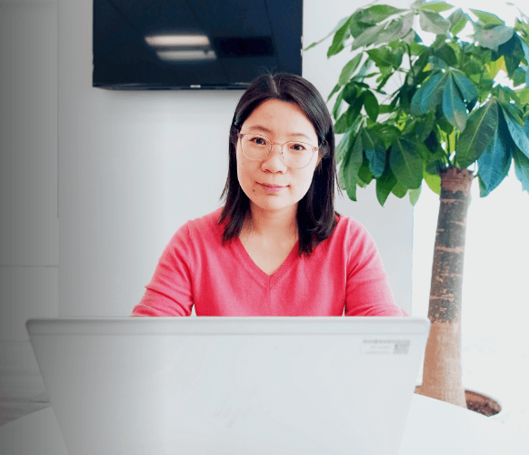 A woman is sitting in front of a computer, next to her, there is a plant and behind her, a TV set.   The woman has shoulder-length hair, she is wearing glasses a long-sleeved blouse.