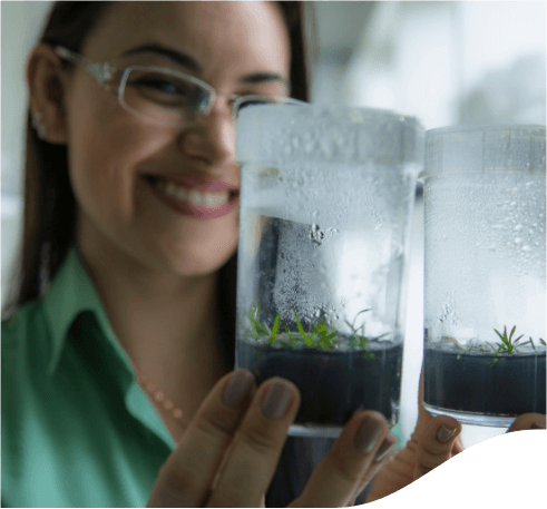 A smiling woman holding a glass vessel with soil and plants inside. She is wearing a light green shirt and eyeglasses.