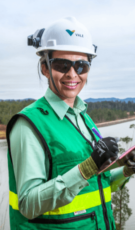 Vale female employee smiling in an operational space. She is wearing a light green shirt a green vest, gloves shirt, goggles and a white helmet the company logo. She is holding a clipboard in one hand.