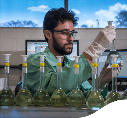 A man inside a laboratory with several glass vessels in front of him. He is wearing goggles and gloves. He is holding one of the glass vessels in his hands, and in the background, are computers.