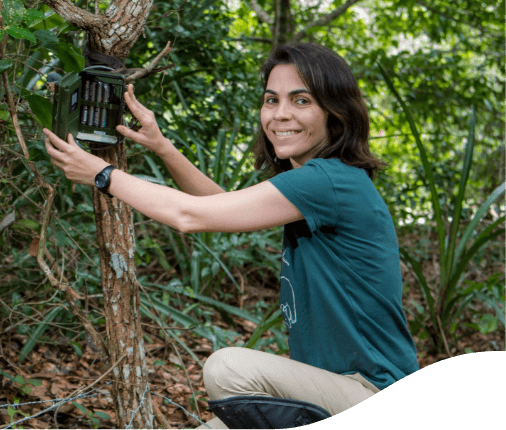 Woman crouching in a space with several trees. She is putting a device on the trunk of one of these trees and looks for the photo smiling. Her hair is down and she is wearing a green T-shirt.