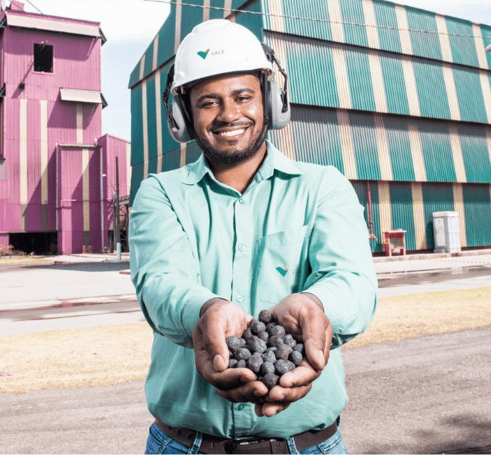 A man smiling a space operational space. He is wearing a light green shirt, ear muffs and a white helmet with Vale logo. He is holding pieces of rock in his hands.