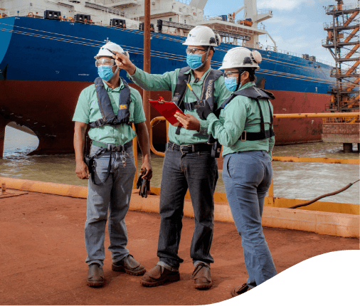 Empregados da Vale parados lado a lado em um porto da empresa. Todos usam uniformes com calças cinzas e camisas verdes, máscara facial, óculos de proteção e capacete branco.