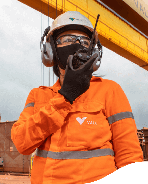 Female employee in an operational area. She is wearing orange uniform with white Vale logo, face mask, goggles, ear muffs and white helmet with Vale logo