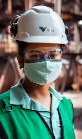 Foto do busto para cima de uma empregada da Vale em uma área operacional. Ela usa camisa verde clara, colete em um tom mais escuro de verde, máscara facial, óculos de proteção e capacete branco com logotipo da Vale.