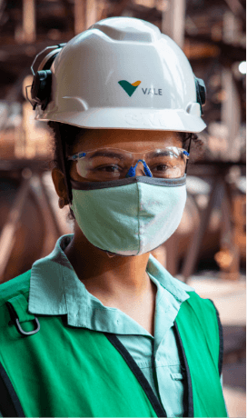 Photo of the chest of Vale female employee in an operational area. She is wearing a light green shirt, a darker green vest,  a face mask, goggles and a white helmet with Vale logo.