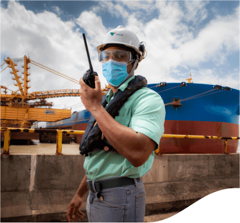 Vale employee standing with a radio communicator in his hands in an outdoor operational area of the company. He is wearing gray pants, a green shirt, a face mask, goggles and a white helmet with Vale logo.