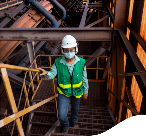 Vale employee climbing the stairs of a metal structure inside an operational area. The employee is wearing jeans, a light green shirt, a darker green vest, a face mask, goggles, ear muffs and a white helmet the company logo.