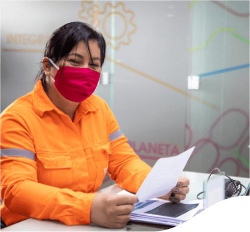 A woman sitting at an office desk. She is holding a sheet of paper in her hands and there are other sheets on the table. The woman is wearing her hair back, orange uniform with gray details and a red face mask.