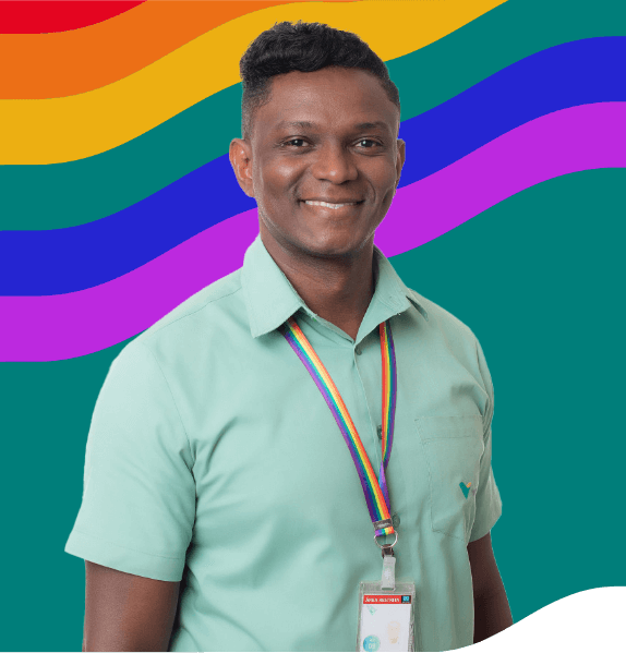 Homem negro sorrindo e usando uma camisa verde clara com logotipo da Vale, além de um crachá com as cores do arco-íris. Atrás dele há uma ilustração da bandeira do arco-íris.