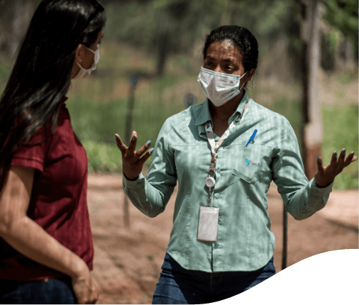 Two women talking in a space with trees. One of them has her hair down and is wearing a red blouse and a face mask. The other has her hair tied back and is wearing a light green shirt, a badge, a face mask and is gesturing with her hands.