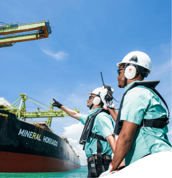 Two Vale employees facing a ship in an operational area. The two are wearing green shirts, protection equipment fixed to the body, goggles, ear muffs, and white helmets.