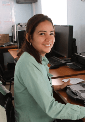 Photo of a smiling woman sitting on a chair working on a computer in an office with a computer in the background. She has shoulder-length dark hair and is wearing a uniform, a green button-up blouse.