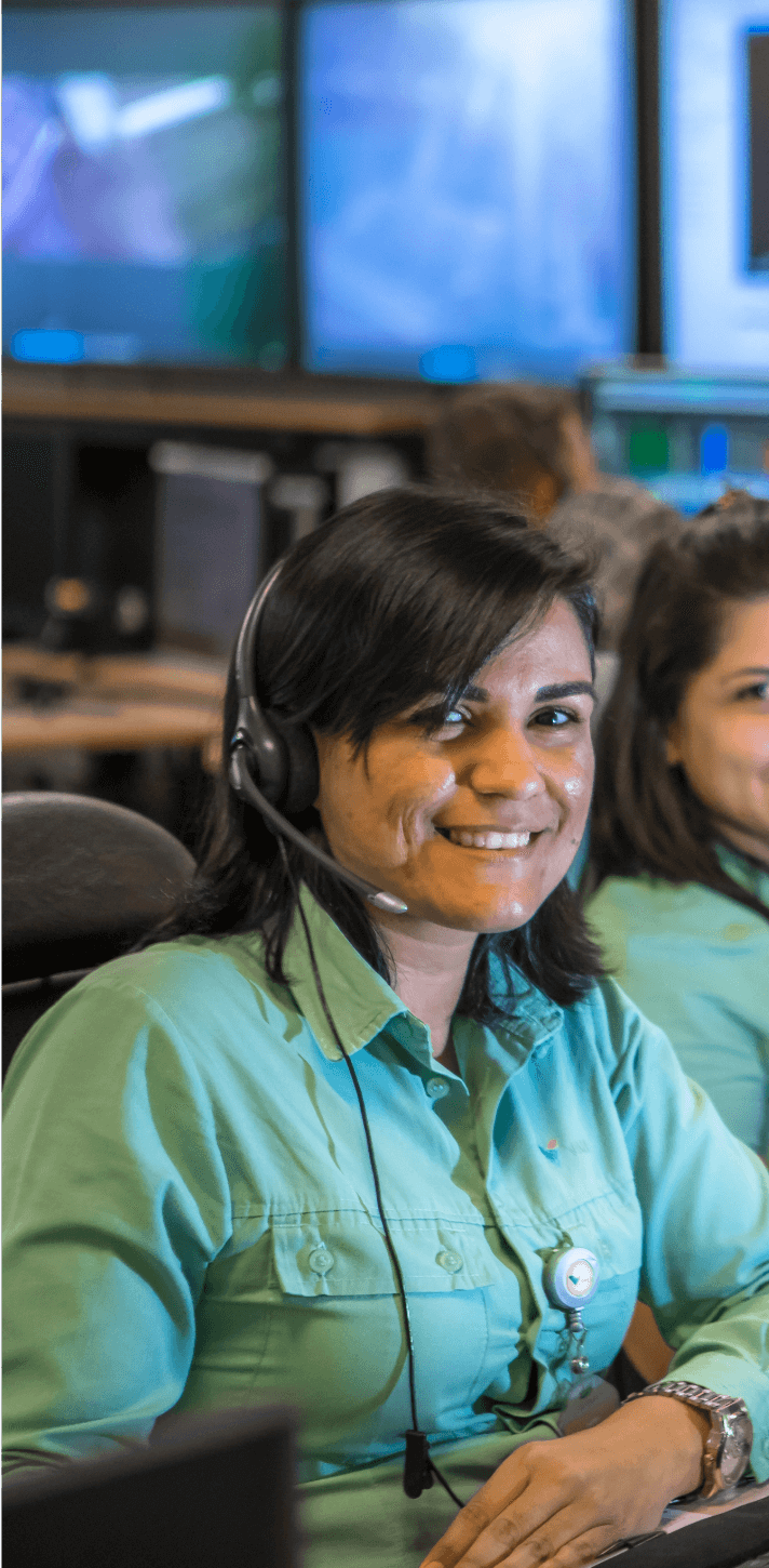 Photo of a woman sitting in an office smiling with a headphone. She has shoulder-length dark hair, bangs to the side, and is wearing a Vale green button-up shirt.