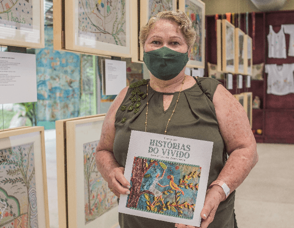 Photo of a woman with short blond hair wearing a tank top and a face mask holding a book in front of her. Behind her, there are several paintings on the wall.