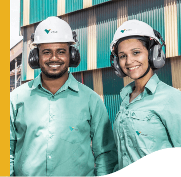 A man and a woman side by side in an operations space. The two are smiling and wearing light green shirts with Vale logo, ear muffs and white helmets also with the company logo.