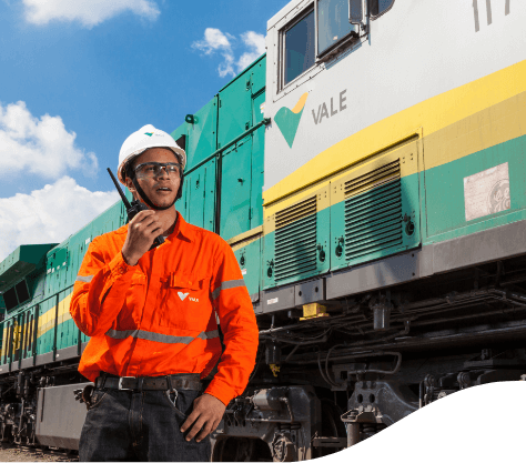 Empregado Vale, utilizando um rádio transmissor. Ele está na frente de um trem, usa um uniforme laranja, capacete e óculos de proteção.