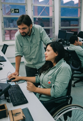 Em um escritório, uma mulher de cadeira de rodas está mexendo em um computador, enquanto um homem está de pé ao lado dela, observando.