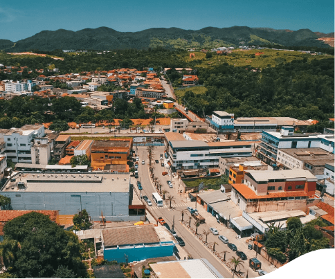 Aerial image of a city. In the foreground, there is an avenue, houses and shops, and in the background, it is possible to see a lot of vegetation and mountainous areas.